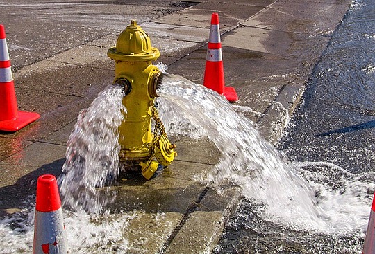 An image of a yellow fire hydrant flowing water on the side of the street, surrounded by orange safety cones.